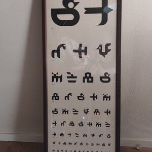 May include: A black and white eye chart for children in a brown frame. The chart features a series of abstract shapes and symbols, with the text "Tavola optometrica per bambini" at the top. The text below reads "sec. Prof. Rizzini distanza 5 metri".