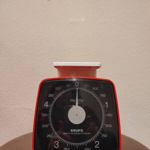 Puede incluir: Una b&aacute;scula de cocina roja y negra con un dial blanco y una cara negra. La b&aacute;scula marca 5 kg 25g. La b&aacute;scula est&aacute; fabricada por Krups y est&aacute; fabricada en la Rep&uacute;blica de Irlanda.