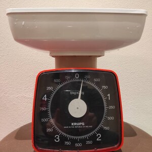 Puede incluir: Una b&aacute;scula de cocina roja y negra con un cuenco blanco en la parte superior. La b&aacute;scula tiene un dial con n&uacute;meros del 0 al 750 y una aguja que apunta a 0. La b&aacute;scula indica "5kg x 25g" y "KRUPS MADE IN THE REPUBLIC OF IRELAND".
