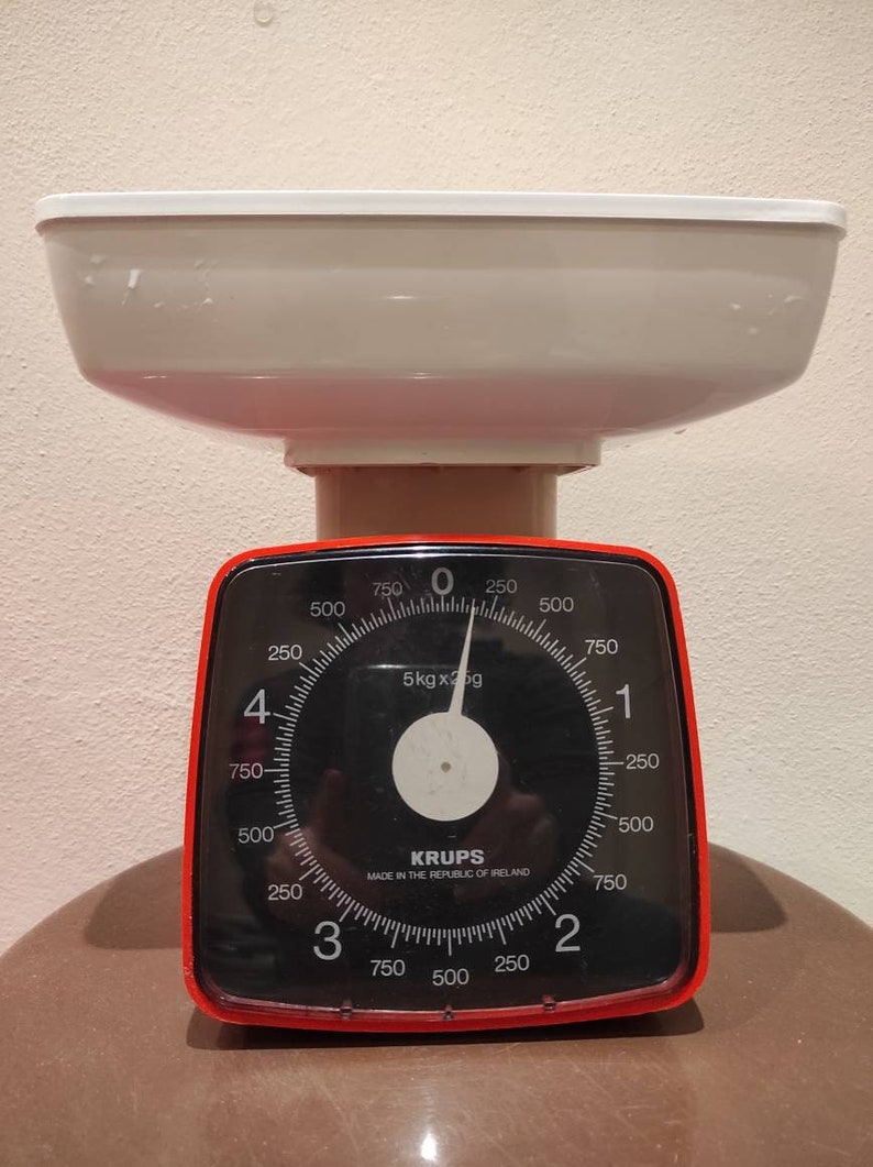 Puede incluir: Una b&aacute;scula de cocina roja y negra con un cuenco blanco en la parte superior. La b&aacute;scula tiene un dial con n&uacute;meros del 0 al 750 y una aguja que apunta a 0. La b&aacute;scula indica "5kg x 25g" y "KRUPS MADE IN THE REPUBLIC OF IRELAND".