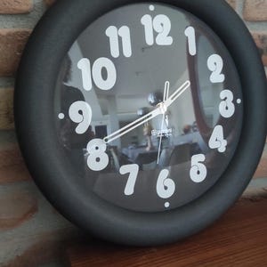 Könnte beinhalten: Eine schwarze runde Wanduhr mit weißen Zahlen und einem silbernen Metallrahmen. Das Zifferblatt der Uhr ist reflektierend und zeigt die Reflexion eines Raumes.