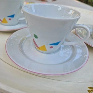Könnte beinhalten: Ein Set aus weißen Keramik-Teetassen und Untertassen mit einem geometrischen Design mit rosa, gelben, blauen und grünen Formen. Die Tassen haben eine abgerundete Form und einen Henkel auf der rechten Seite.