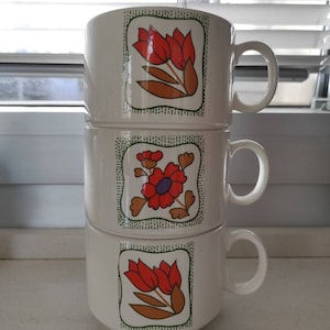 Può includere: Tre tazze in ceramica bianca impilate l'una sull'altra. Ogni tazza ha un diverso disegno floreale con fiori rossi, arancioni, verdi e viola. Le tazze hanno uno sfondo bianco con un motivo a pois verde e bianco.