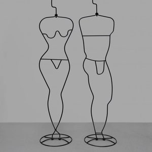 Op de afbeelding: Twee zwarte metalen mannequins, een met een vrouwelijke vorm en een met een mannelijke vorm, staan op een grijze achtergrond. De mannequins zijn gemaakt van dunne draad en hebben een eenvoudig, minimalistisch ontwerp.