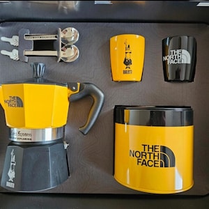May include: A yellow and black coffee set, including a stovetop espresso maker, two small cups, a container, and two spoons. The set features "The North Face" branding. The espresso maker has a black handle and base.