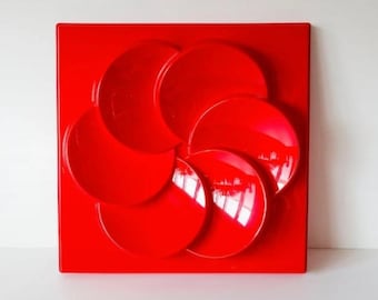 May include: A striking red wall art piece. It features a square base with six overlapping, glossy, circular shapes. The design creates a three-dimensional effect, reflecting light. This artwork is a bold statement piece for any room.