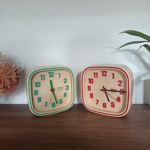 Op de afbeelding: Twee vierkante wekkers met crèmekleurige wijzerplaten. Eén heeft een groene rand en wijzers, de andere een rode rand en wijzers. Beide klokken hebben zwarte cijfers en het woord "bino quartz".