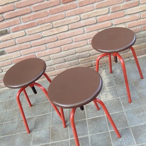 May include: Three brown metal stools with red legs. The stools are arranged in a row on a tiled floor.