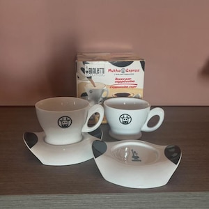 Op de afbeelding: Twee witte keramische cappuccinokopjes met bijpassende schoteltjes voorzien van een zwart koeienkoplogo. De schoteltjes hebben zwarte koeienvlekken aan de zijkanten. De set staat voor een Bialetti Mukka Express doos met producttekst.