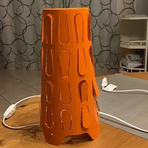 May include: Orange table lamp with a unique cut-out design. The lamp is on a wooden table and has a white cord.