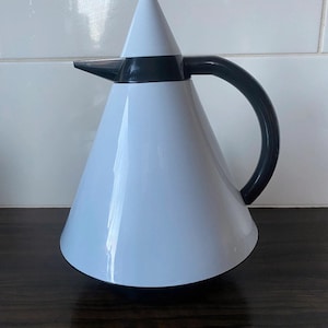 Può includere: Caraffa termica bianca e nera con corpo a forma di cono e manico nero. La caraffa ha un coperchio nero con un piccolo pomo nero in cima.