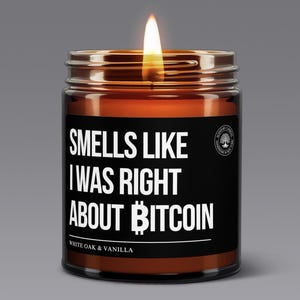 May include: A brown glass candle with a black label that reads "Smells Like I Was Right About Bitcoin". The candle is lit and the flame is visible. The label also includes the text "White Oak & Vanilla".