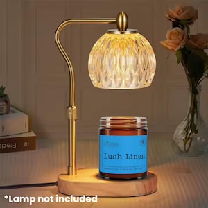 May include: A gold-toned, arched lamp with a textured glass shade illuminates a wooden base. A jar of "Lush Linen" scented candle sits on the base. The text "*Lamp not included" is at the bottom.