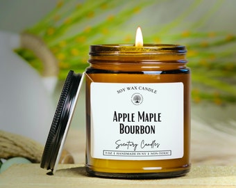 May include: A lit soy wax candle in a brown glass jar with a black lid. The label reads "Apple Maple Bourbon" and "Scentury Candles." The candle is 9 oz and handmade in NY. The background is blurred with green and white.