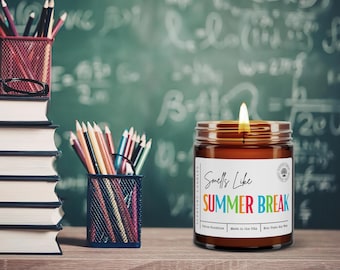 May include: A lit candle in a glass jar with a label that reads "Smells Like SUMMER BREAK" in colorful letters. The candle sits on a wooden surface with pencils, books, and a chalkboard in the background.