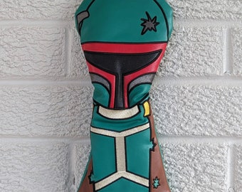 May include: A green, red, and brown golf club head cover with a Star Wars Boba Fett design. The cover has a brown leather-like material on the bottom and a green leather-like material on the top. The cover has a red and black face mask design with a white star on the top. The text "fairway" is printed in red below the cover.