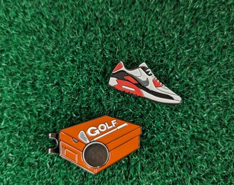 May include: Two enamel pins featuring a golf shoe and a golf shoe box with the word "GOLF" on it. The pins are on a green background.