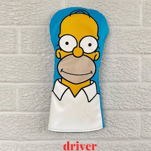 May include: A yellow golf club head cover with a cartoon image of Homer Simpson from the Simpsons television show. The cover is blue on the top and white on the bottom. The text "driver" is printed in red below the cover.