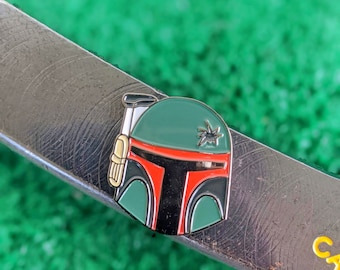 May include: A green, red, and black enamel pin featuring a Star Wars Boba Fett character. The pin is on a silver surface.