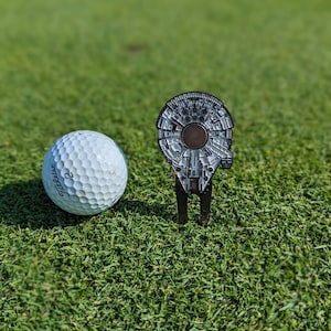 May include: A white golf ball with the text "Titleist 2" on it, next to a black golf ball marker shaped like the Millennium Falcon from Star Wars.