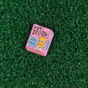 May include: A pink and blue enamel pin featuring a cartoon character with yellow skin, a blue shirt, and a yellow tie. The character is holding a blue and white box with the text "ミスター スモール" in Japanese. The pin is on a green background.