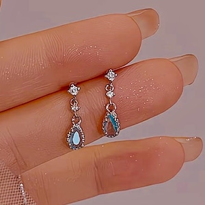 Puede incluir: Pendientes colgantes plateados con una piedra preciosa azul en forma de lágrima y tres pequeños cristales transparentes. Los pendientes están sostenidos por una mano, mostrando el delicado diseño y brillo de la joyería.