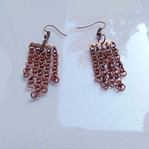 Puede incluir: Un par de pendientes de cadena de cobre con un diseño colgante. Los pendientes están hechos de múltiples eslabones pequeños de cadena de cobre.