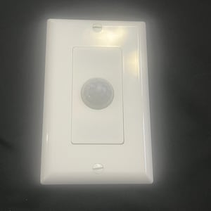 Puede incluir: Un interruptor de luz con sensor de movimiento blanco con una placa frontal rectangular. El interruptor tiene una sección rectangular más pequeña en el centro con un sensor circular. Dos tornillos aseguran la placa frontal a la pared.