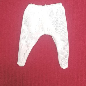 Peut inclure: Un pantalon en tricot blanc à mailles ajourées, avec un bord festonné à la taille. Le pantalon est fait d'un tissu délicat et aéré, et est présenté sur un fond rouge. Le pantalon semble être pour un bébé ou un tout-petit.