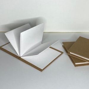 May include: A handmade book with white pages and a brown cardboard cover, open to display the pages. Several similar books are stacked nearby. The book is on a white surface.