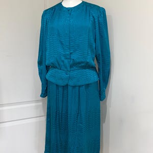 May include: A teal two-piece dress set featuring a long-sleeved button-down blouse with a round neckline and a matching knee-length skirt. The fabric has a subtle, repeating textured pattern throughout, and the blouse includes a fitted waist detail.