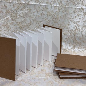 Może przedstawiać: Wyświetlanych jest kilka małych, pustych notatników z brązowymi okładkami i białymi stronami. Jeden jest otwarty, pokazując rozłożone strony. Inny jest otwarty, a także widoczny jest stos trzech zamkniętych notatników.