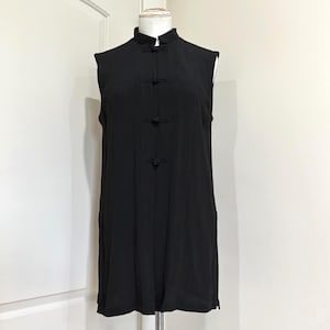 May include: A sleeveless black tunic featuring a high mandarin collar and a front placket secured with four fabric-covered knot buttons. The garment has a straight, relaxed silhouette with a subtle vertical texture throughout the fabric.