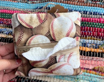 Puede incluir: Una bolsa de tela cuadrada con un patrón repetitivo de pelotas de béisbol con costuras rojas sobre un fondo beige. La bolsa incluye una abertura horizontal con cremallera en el centro, de la cual sobresale una toallita de tela blanca y suave.