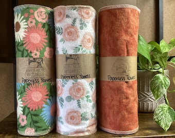 Op de afbeelding: Drie cilindrische rollen herbruikbare stoffen handdoeken, omwikkeld met bruine papieren banden met het label Hippie Mom Reusables Paperless Towels. De linkerrol heeft een groene bloemenprint, de middelste rol een witte bloemenprint en de rechterrol is effen roestoranje.