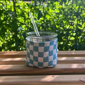 May include: A clear glass jar with a blue and white chequered cosy and a clear glass straw. The jar sits on a wooden surface with a green leafy background. The cosy is wrapped around the jar's exterior.