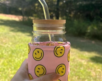 May include: A clear glass mason jar with a bamboo lid and a bent glass straw. The jar is encased in a light pink fabric sleeve featuring a repeating pattern of yellow smiley faces with black outlines and features, providing a secure grip.