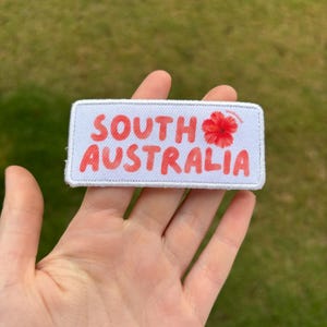 May include: A rectangular white embroidered patch with a stitched border. The text SOUTH AUSTRALIA is printed in a bold, coral-pink font, accompanied by a small, detailed illustration of a red hibiscus flower positioned to the right of the text.