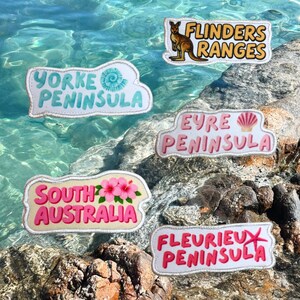 May include: Five embroidered patches featuring South Australian locations. The patches display the text Flinders Ranges with a kangaroo, Yorke Peninsula with a shell, Eyre Peninsula with a scallop, South Australia with hibiscus flowers, and Fleurieu Peninsula with a starfish.
