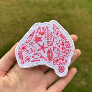 May include: A white fabric patch shaped like the continent of Australia, featuring intricate red line art illustrations of seashells, a sea turtle, a starfish, a seahorse, and tropical flowers, with the word sunbound printed in small red text.