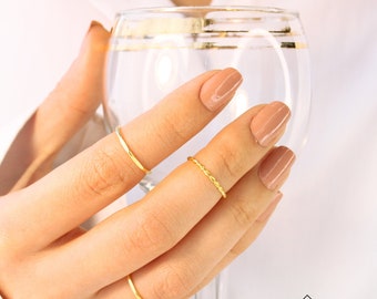 May include: A hand wearing three thin gold rings on the fingers. The hand is holding a glass of water. The rings are simple and delicate.