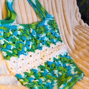 Puede incluir: Bolso tote de crochet hecho a mano en una vibrante mezcla de verde, azul y blanco. El bolso presenta un diseño de granny square y un asa resistente. La construcción de tejido abierto es visible, mostrando la detallada artesanía.