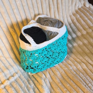 May include: A turquoise, white, and black crochet basket containing grey and black yarn. The basket features a scalloped edge and an open, textured weave. It rests on a soft, cream-coloured blanket. The basket is ideal for storing yarn.