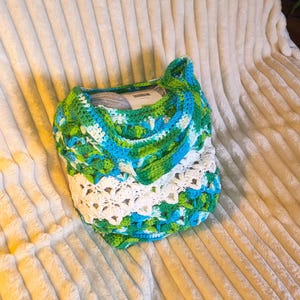 May include: A handmade crochet bag in shades of blue, green, and white. The bag features an openwork design and contains a ball of grey yarn. The bag is on a soft, white, textured surface.