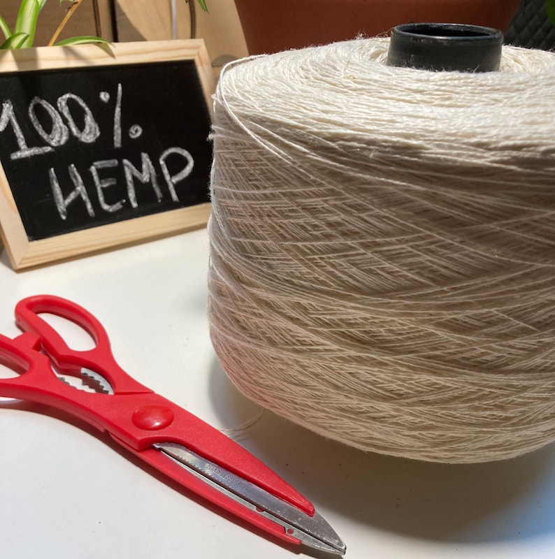 May include: A spool of natural beige hemp yarn with a black plastic spool holder. The yarn is 100% hemp. A red pair of scissors is on a white surface.