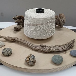 May include: A spool of natural white yarn on a wooden platform with driftwood, stones, and shells.