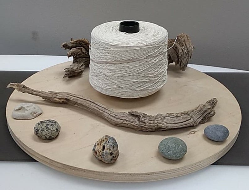 May include: A spool of natural white yarn on a wooden platform with driftwood, stones, and shells.
