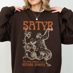 Könnte beinhalten: Ein dunkelbraunes Sweatshirt mit dem Wort "SATYR" in orangefarbenen Buchstaben. Darunter eine Illustration einer mythologischen Figur, die Panflöte spielt, mit dem Text "PAN PIPES CONCERT BY THE NATURE SPIRITS".