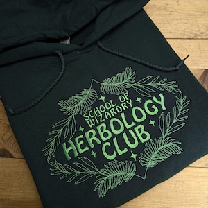 May include: Dark teal hoodie with a drawstring hood. The front features the text "SCHOOL OF WIZARDRY HERBOLOGY CLUB" in green, surrounded by a green leaf design. The hoodie is folded on a wooden surface.