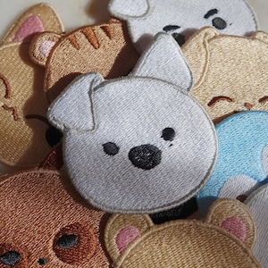 May include: A collection of embroidered patches featuring various animal faces, including dogs, cats, and bears. The patches are in shades of brown, tan, white, and blue, with black and pink accents. The patches are round and have a stitched border.